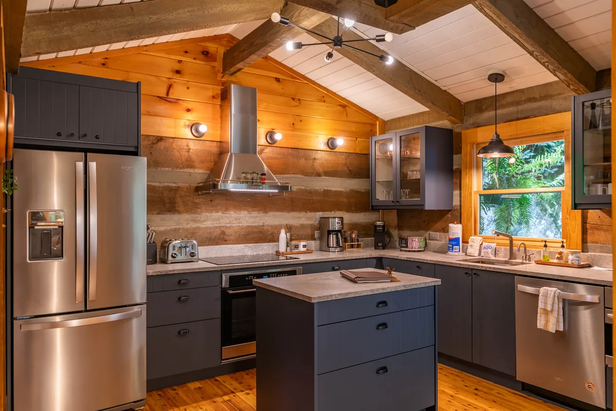 A modern, chef-inspired kitchen is featured here, with a spacious kitchen island, sleek stainless steel appliances, stone countertops, exposed wooden beams, and a large window offering views of the greenery outside. Under-cabinet lighting creates an inviting ambiance.