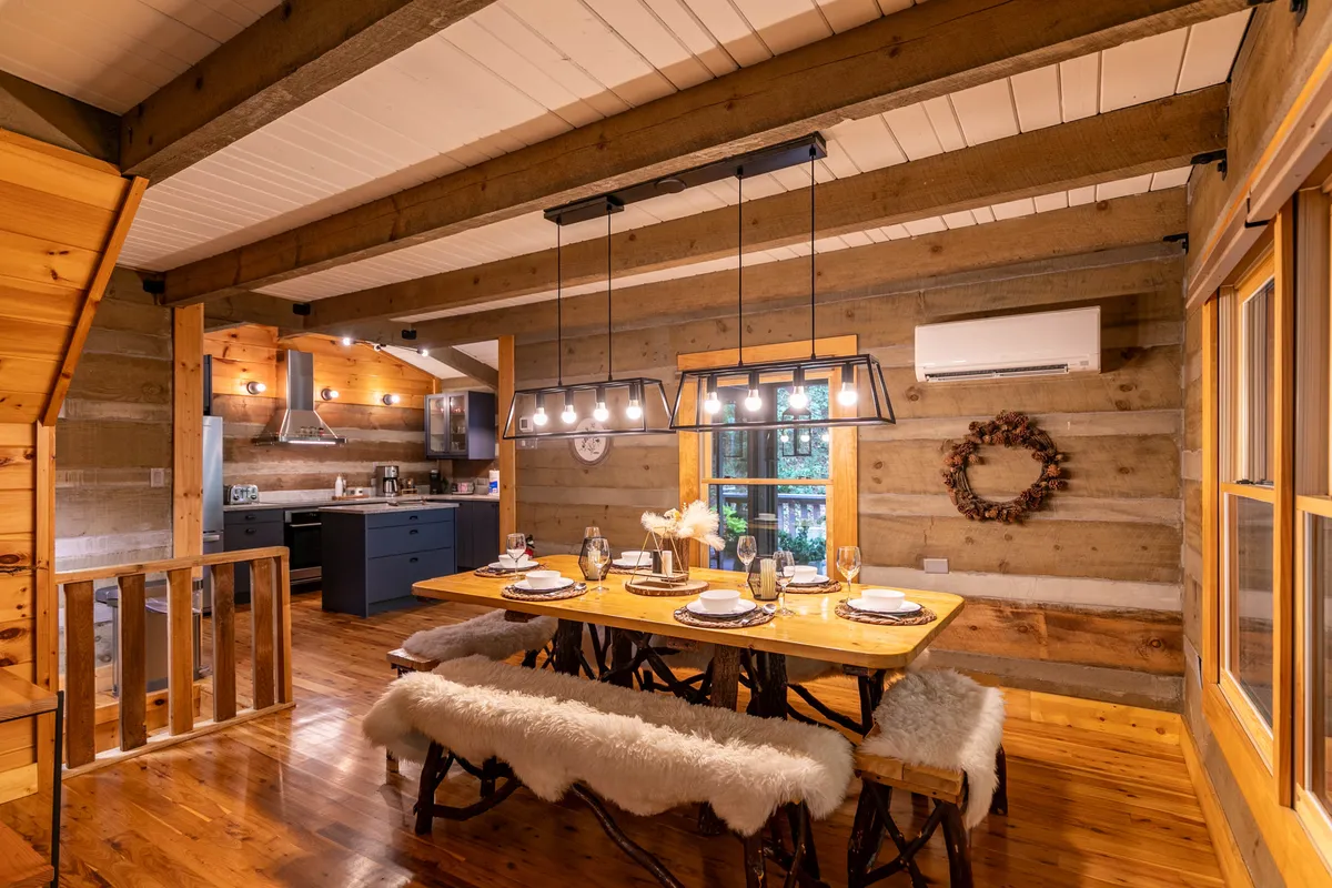This image focuses on the dining area within the open-plan space. The rustic wooden dining table sits beneath a statement light fixture, with exposed beams above and the kitchen area visible in the background, tying together dining and culinary spaces.