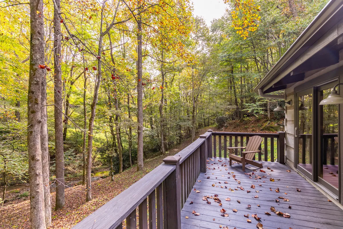 A serene wooden deck surrounded by lush trees and overlooking a gentle stream, with fallen leaves adding a touch of seasonal beauty. The setting feels secluded and peaceful, ideal for couples or small groups wanting to relax, read, or meditate amidst nature.