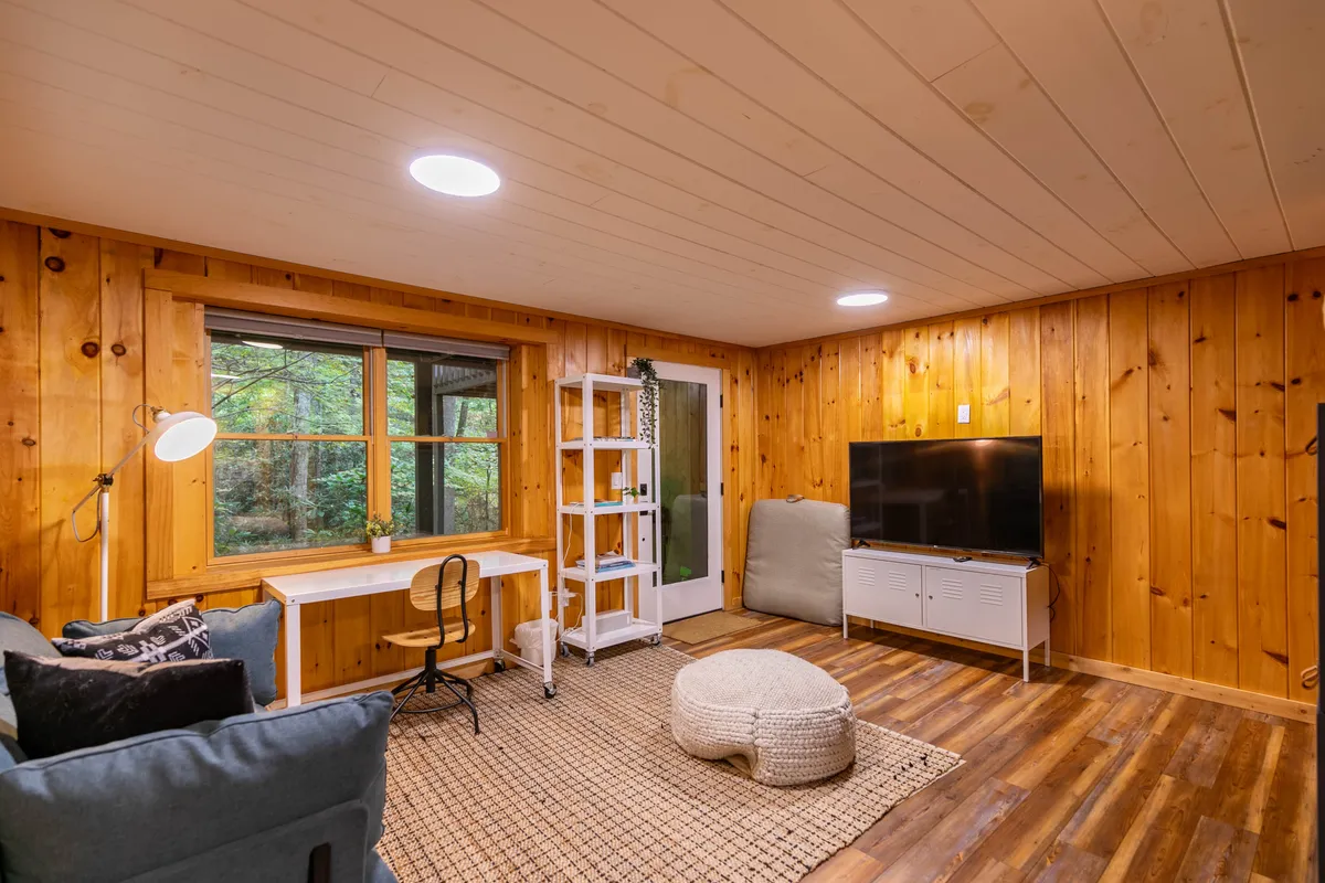 This image features a cozy interior space with wooden walls, a large window, a TV, a desk with a chair, and a bean bag chair. The space looks inviting and comfortable, ideal for relaxing, watching TV, or enjoying quiet downtime.