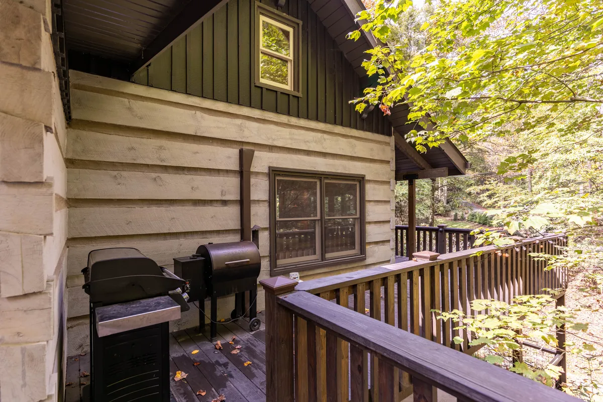 A cozy wooden deck nestled among trees, furnished with a grill and outdoor seating, offering an inviting spot for morning coffee, outdoor meals, or evening stargazing. The enveloping forest backdrop highlights immersive nature experiences.
