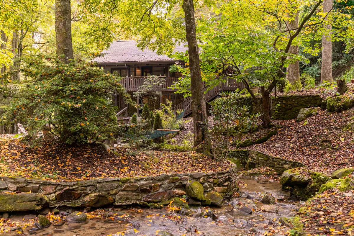 A picturesque view of the house nestled among tall trees, with a gentle stream flowing nearby and a rustic stone wall in the foreground. The tranquil landscape offers a sense of seclusion while inviting exploration of the natural surroundings.