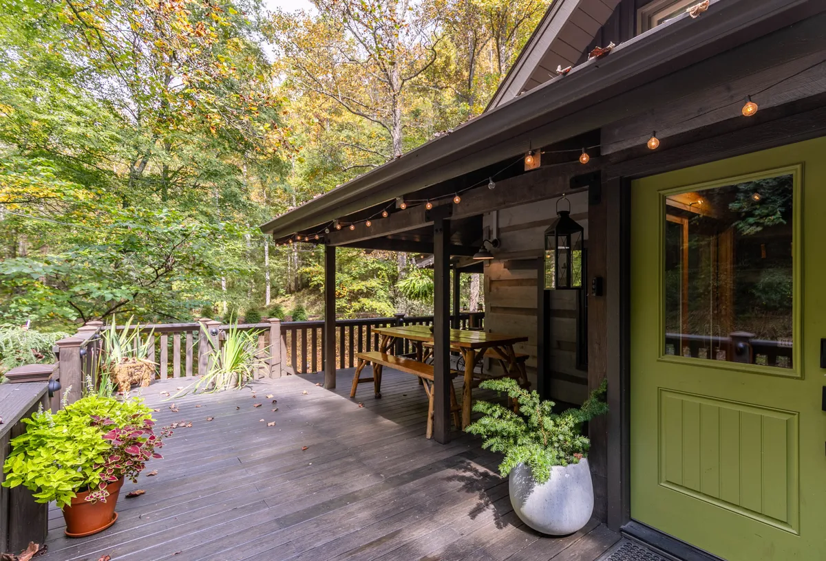 A welcoming exterior deck adorned with string lights, a green front door, wooden deck flooring, a dining table, and potted plants. The space exudes warmth and is clearly designed for outdoor relaxation or shared meals, with an inviting atmosphere perfect for evenings or mornings spent together.