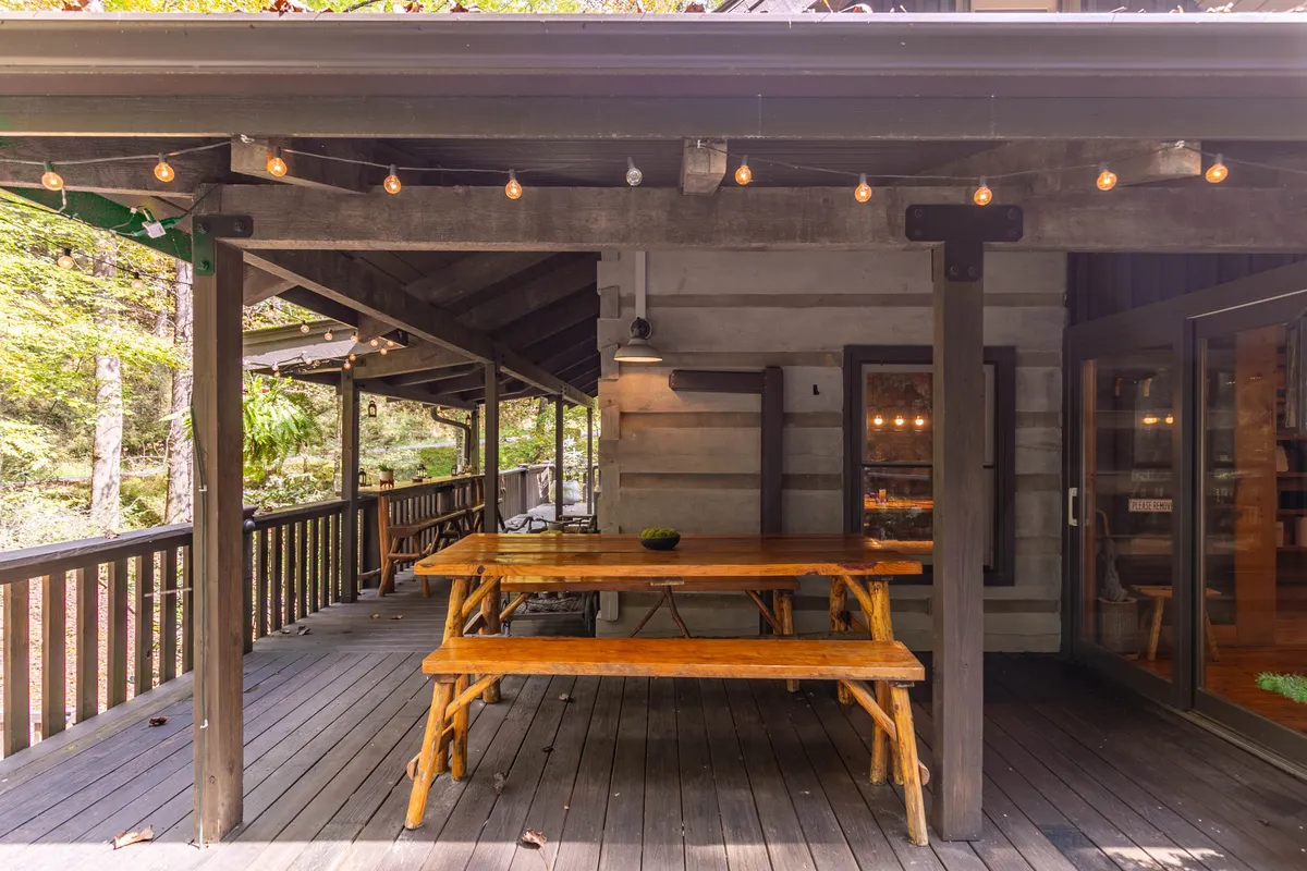 A wooden picnic table sits under string lights amidst lush greenery, creating a casual and welcoming atmosphere for outdoor dining, games, or festive celebrations. The scene is framed by wooden rails and surrounded by nature.