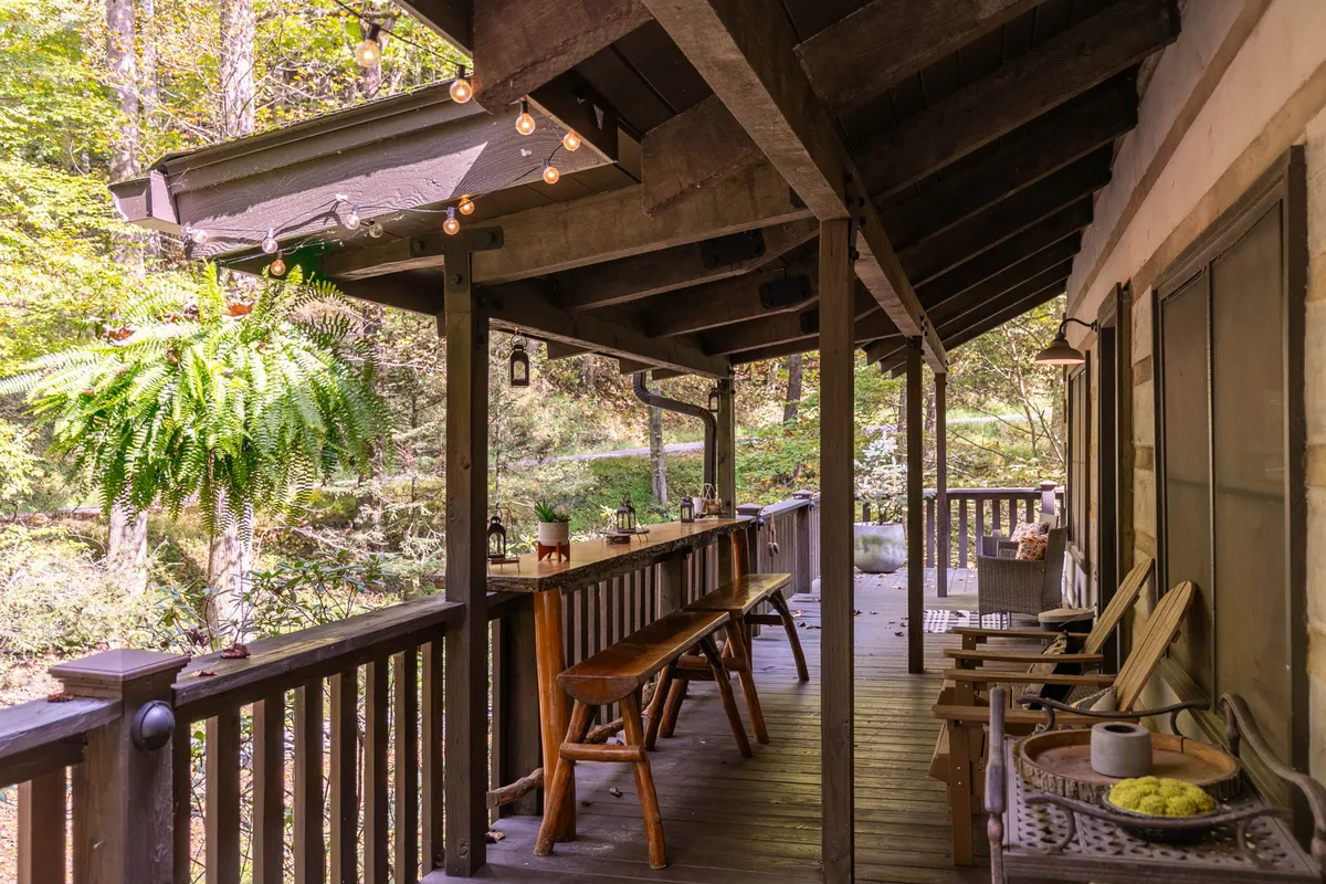 A spacious wooden deck adorned with string lights and hanging ferns, featuring a long table and comfortable seating that invites relaxation and socializing. The deck is surrounded by lush greenery, creating a seamless connection between the home and its natural environment.