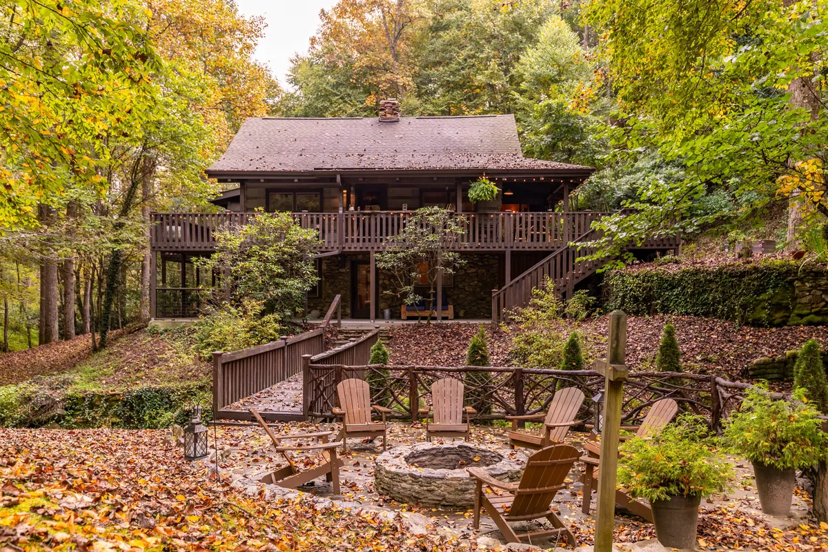 The hero image showcases a charming wooden deck overlooking a crackling fire pit, nestled among tall, lush trees. The stone facade of the updated cabin exudes rustic elegance, while the serene natural surroundings invite relaxation and outdoor gatherings. The setting suggests opportunities for sunset watching, stargazing, and tranquil moments immersed in nature.