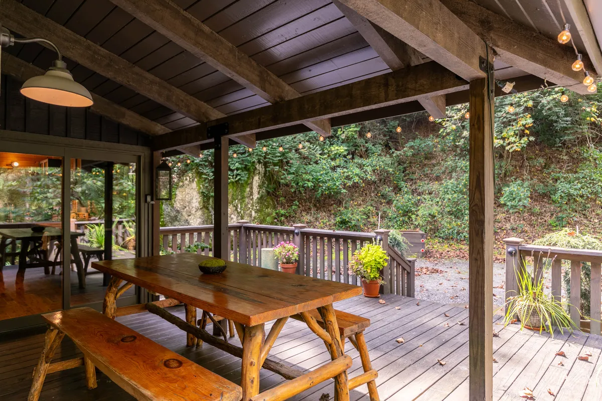 A long wooden dining table sits beneath string lights on a deck surrounded by lush greenery and decorative plants. The outdoor dining setup invites guests to enjoy shared meals, celebrations, or relaxed mornings together in the fresh mountain air.