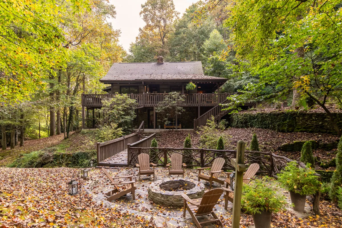 An expansive wooden deck overlooks the surrounding forest, with a prominent fire pit encircled by comfortable seating. A stone path leads through the trees, creating an inviting atmosphere for gathering outdoors. The deck and fire pit area are perfect for group conversations, evening relaxation, or stargazing together.