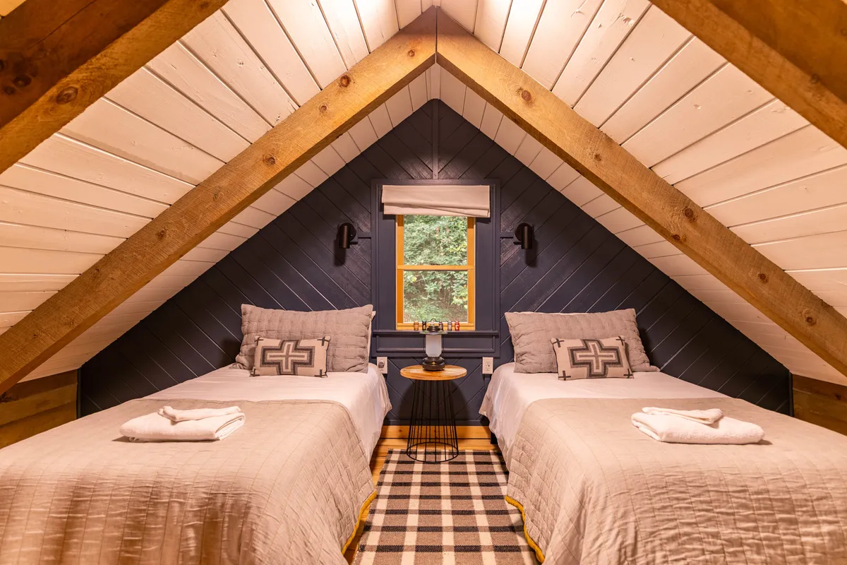 A charming loft area furnished with two twin beds, exposed wooden beams, and a window that lets in natural light. The space is inviting and perfect for restful sleep or cozying up with a book.