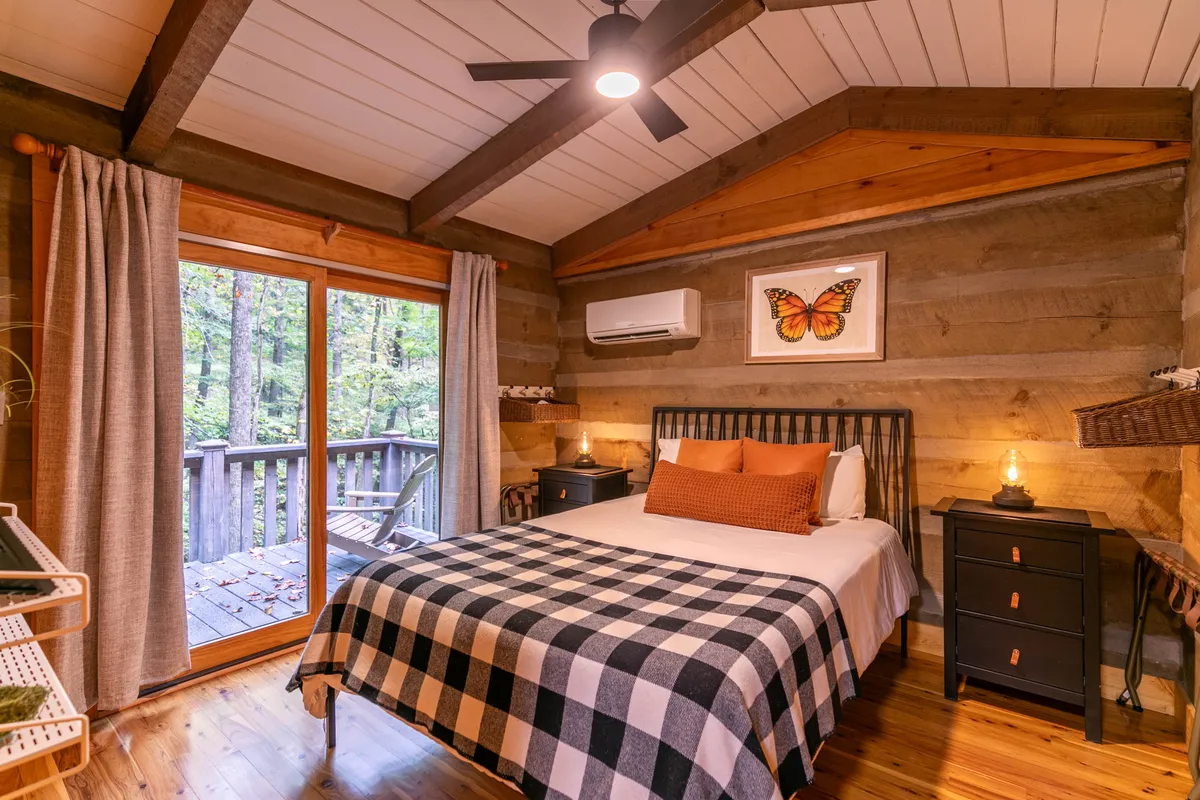 A cozy queen bedroom featuring a bed with a plaid blanket, large windows offering leafy views, butterfly artwork above the headboard, and nightstands with warm lighting. The room exudes welcoming charm and connects guests to the tranquility of nature outside.