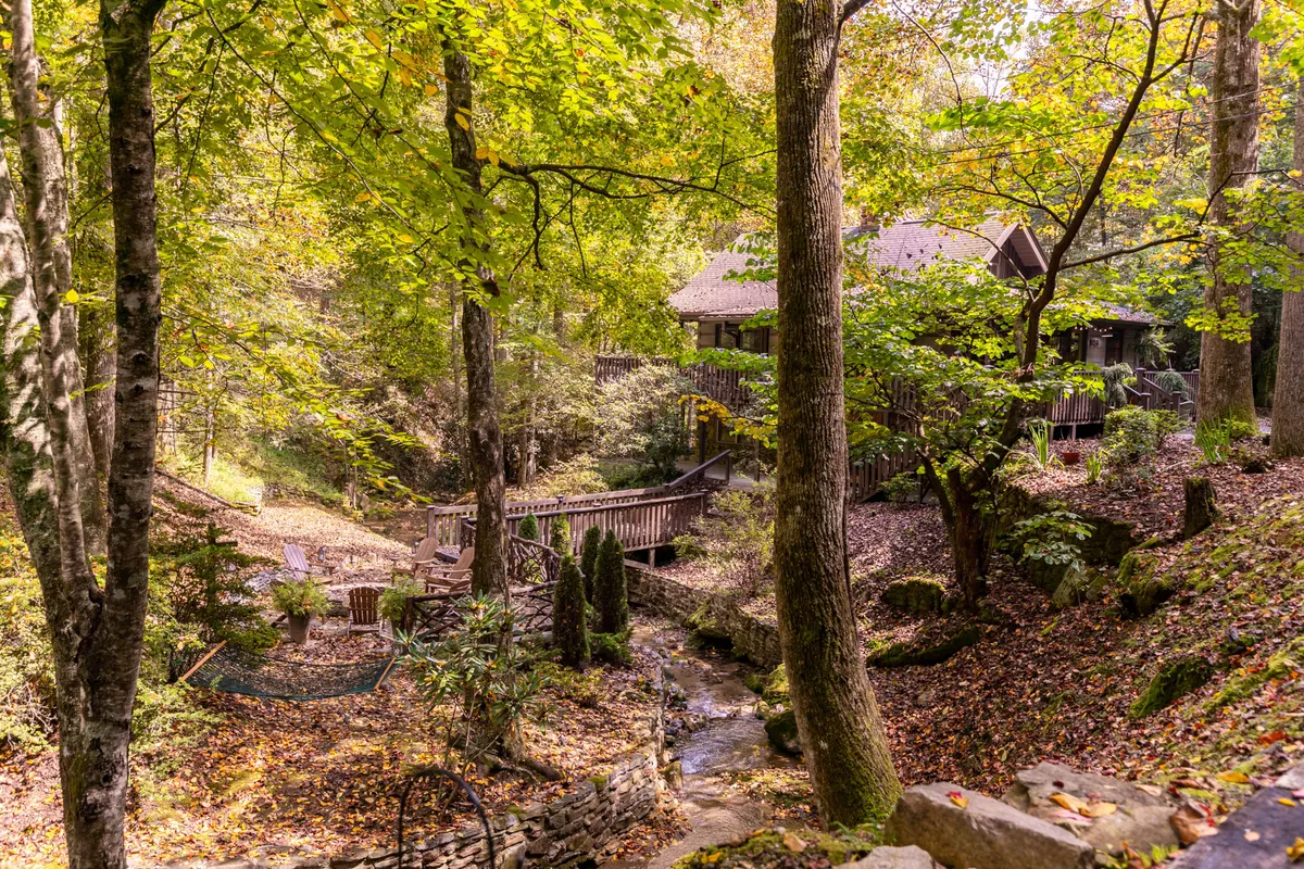 This scenic view highlights the cabin’s relationship with its natural setting, showing the house, a flowing stream, a bridge, and surrounding trees. A seating area by the stream suggests a peaceful spot for relaxation, play, or watching children swim in the creek’s natural pools.