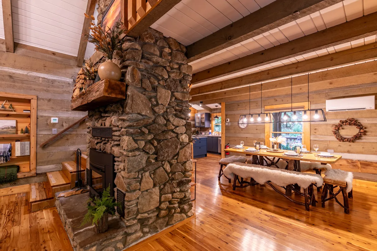 The image presents a view of the living and dining area, with a prominent stone fireplace, rustic wooden beams overhead, and a sizable dining table. The room exudes warmth and is clearly designed for gatherings, with the fireplace as a central feature for both spaces.