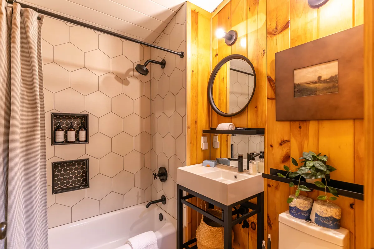 A spa-style, custom-tiled bathroom with a hexagonal tile shower wall, a round mirror, wooden paneling, and a minimalist sink. The ambience is calm and contemporary, with subtle rustic touches.