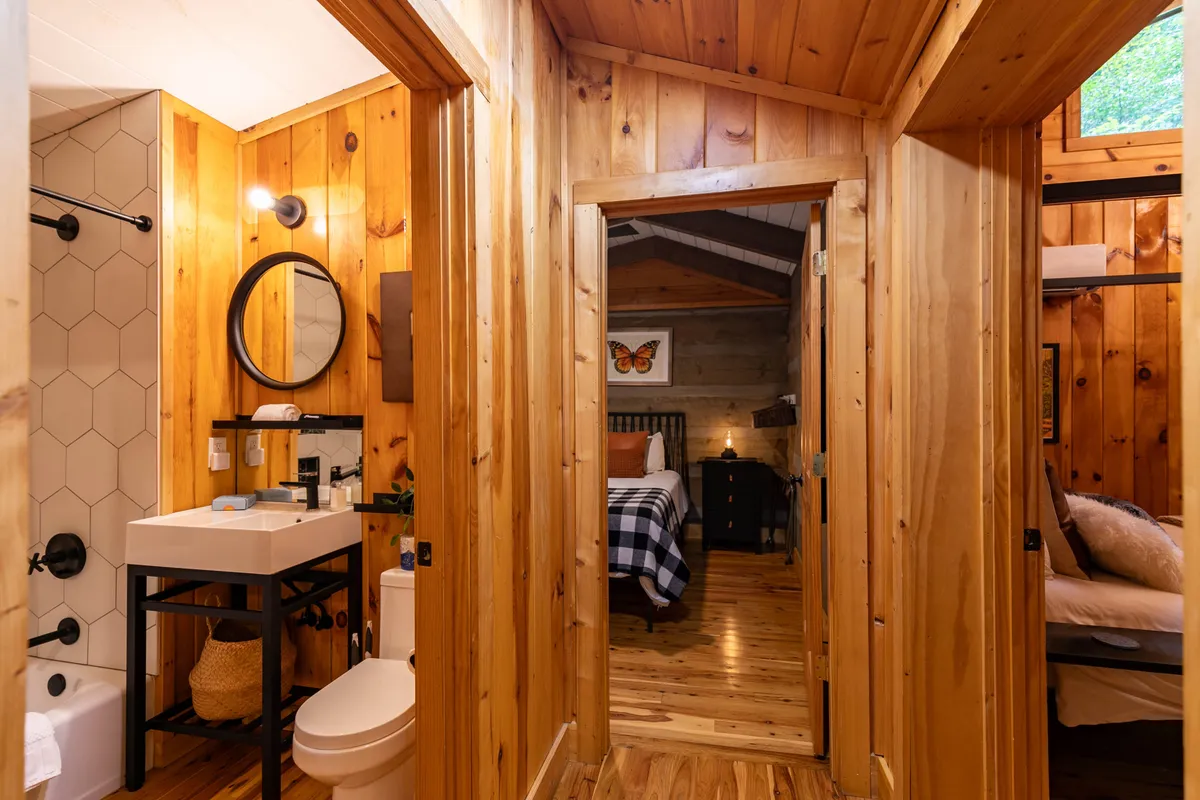 A view of a bathroom sink with a large mirror, wooden walls and ceiling, and a doorway leading into a bedroom. The design blends rustic charm with modern comfort, creating a seamless flow between private retreat and spa-inspired amenities.