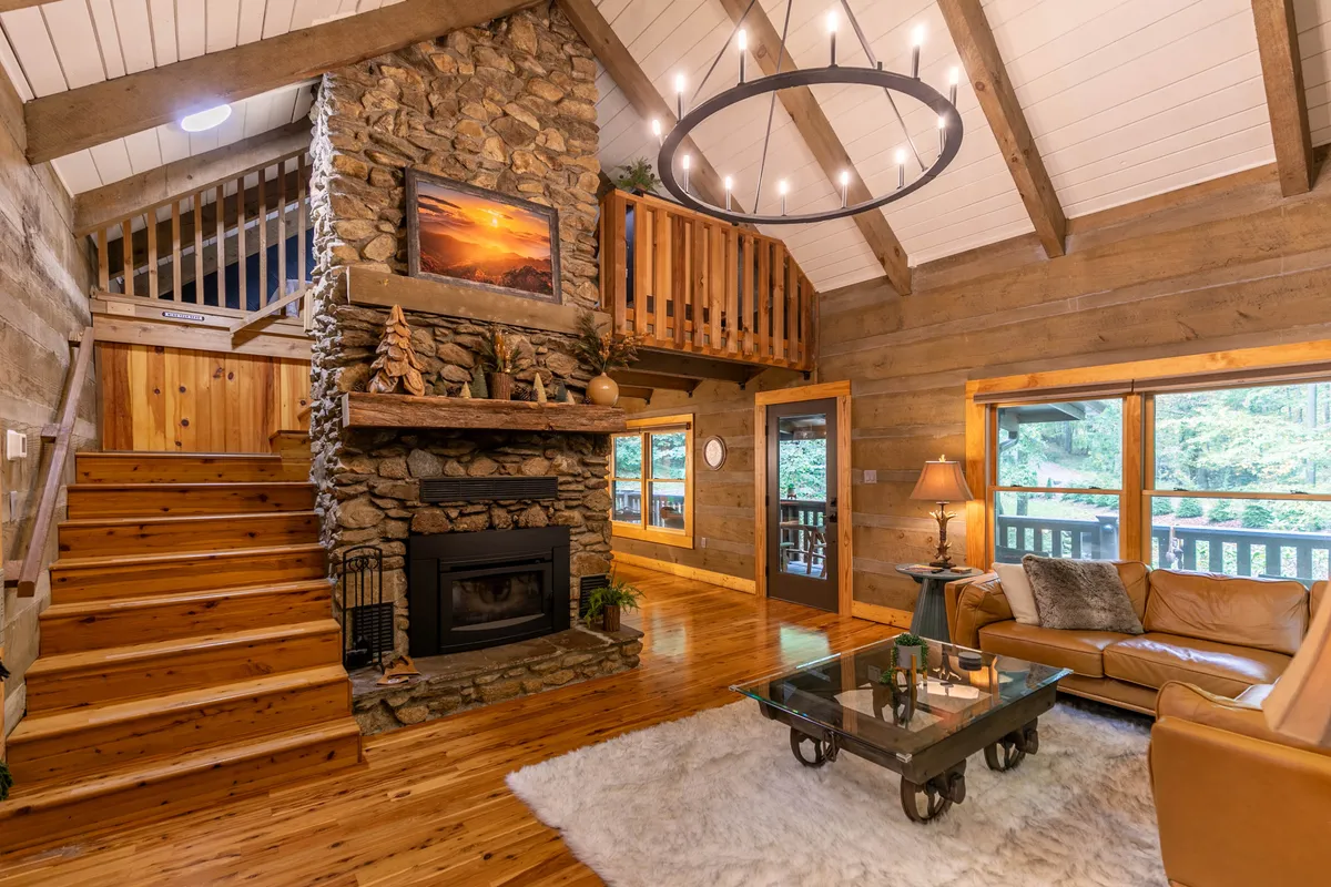 This image captures a grand stone fireplace stretching up two stories, flanked by a wooden staircase and large windows that flood the living area with natural light. A leather sofa anchors the space, while rustic accents like exposed beams and a chandelier add both warmth and elegance.